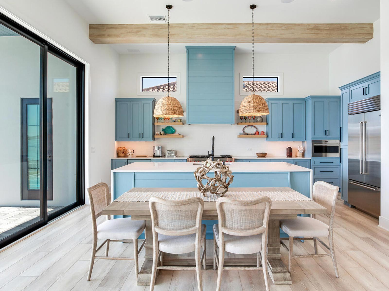 Bright and modern kitchen featuring light blue cabinetry, a spacious island, and a wooden dining table with stylish chairs. Natural light floods the space through large sliding glass doors, highlighting the contemporary design and inviting atmosphere.