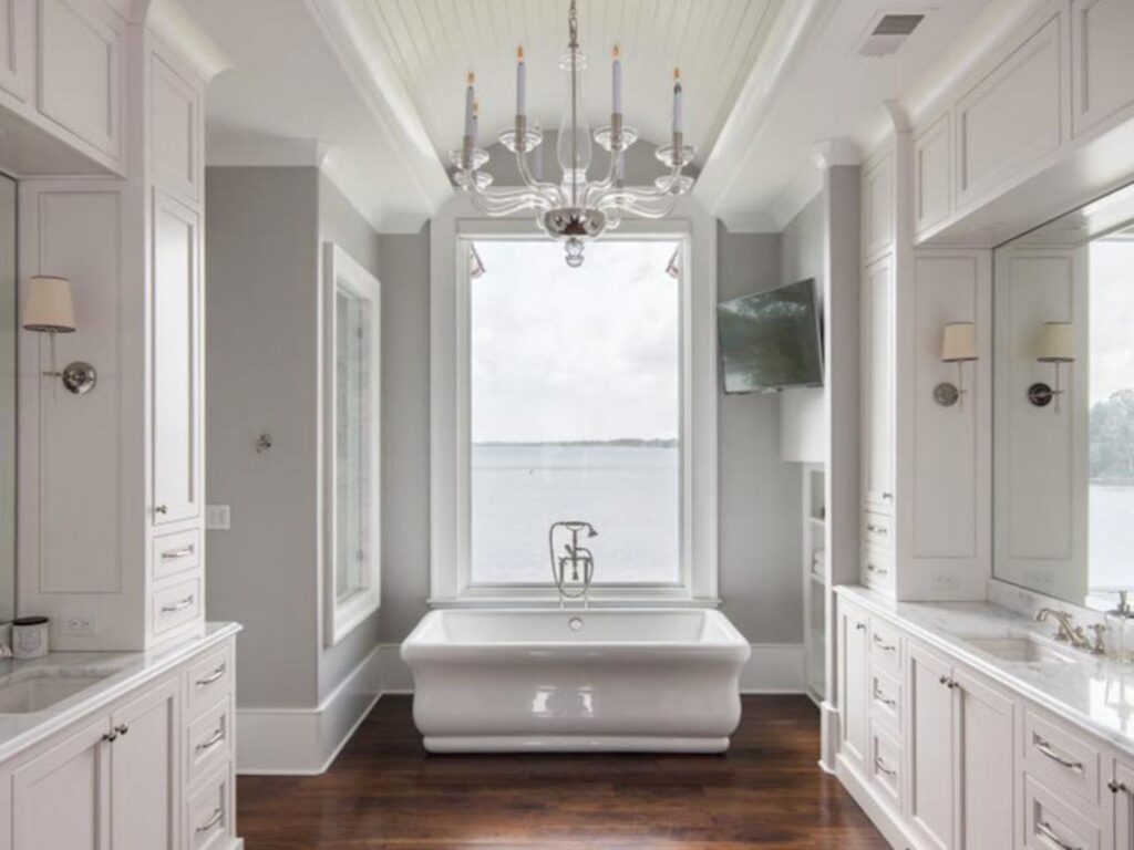 Luxurious modern bathroom featuring a freestanding bathtub, elegant chandelier, and large window with a scenic water view. The space is designed with white cabinetry, marble countertops, and hardwood flooring, creating a serene and sophisticated atmosphere.