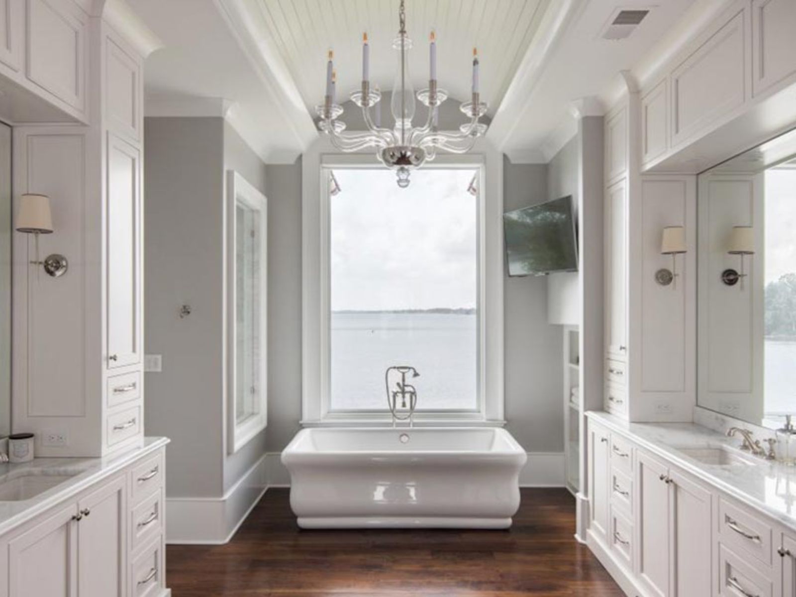 Luxurious modern bathroom featuring a freestanding bathtub, elegant chandelier, and large window with a scenic water view. The space is designed with white cabinetry, marble countertops, and hardwood flooring, creating a serene and sophisticated atmosphere.