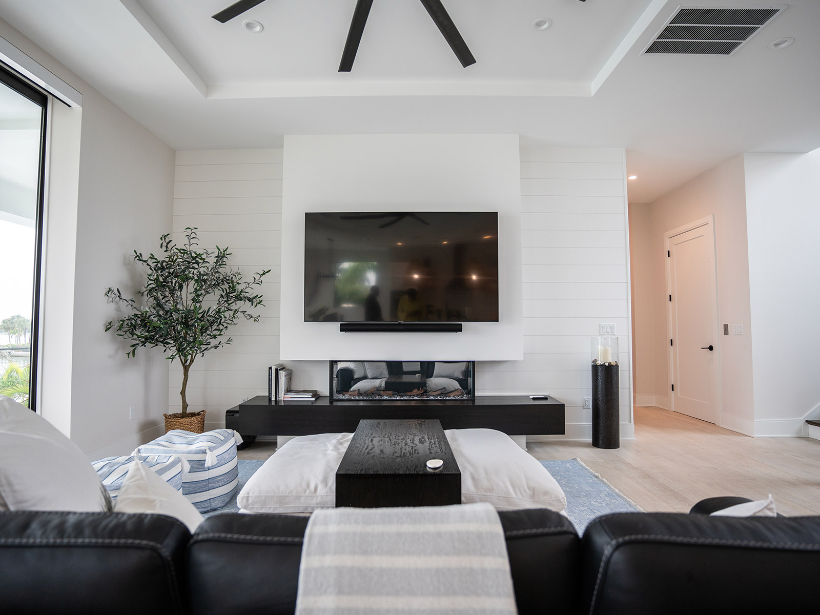 Modern living room featuring a large flat-screen TV mounted on a white shiplap wall, a sleek black media console, and cozy seating with striped bean bags. Natural light floods the space through large windows, complemented by a decorative indoor tree and minimalist decor.