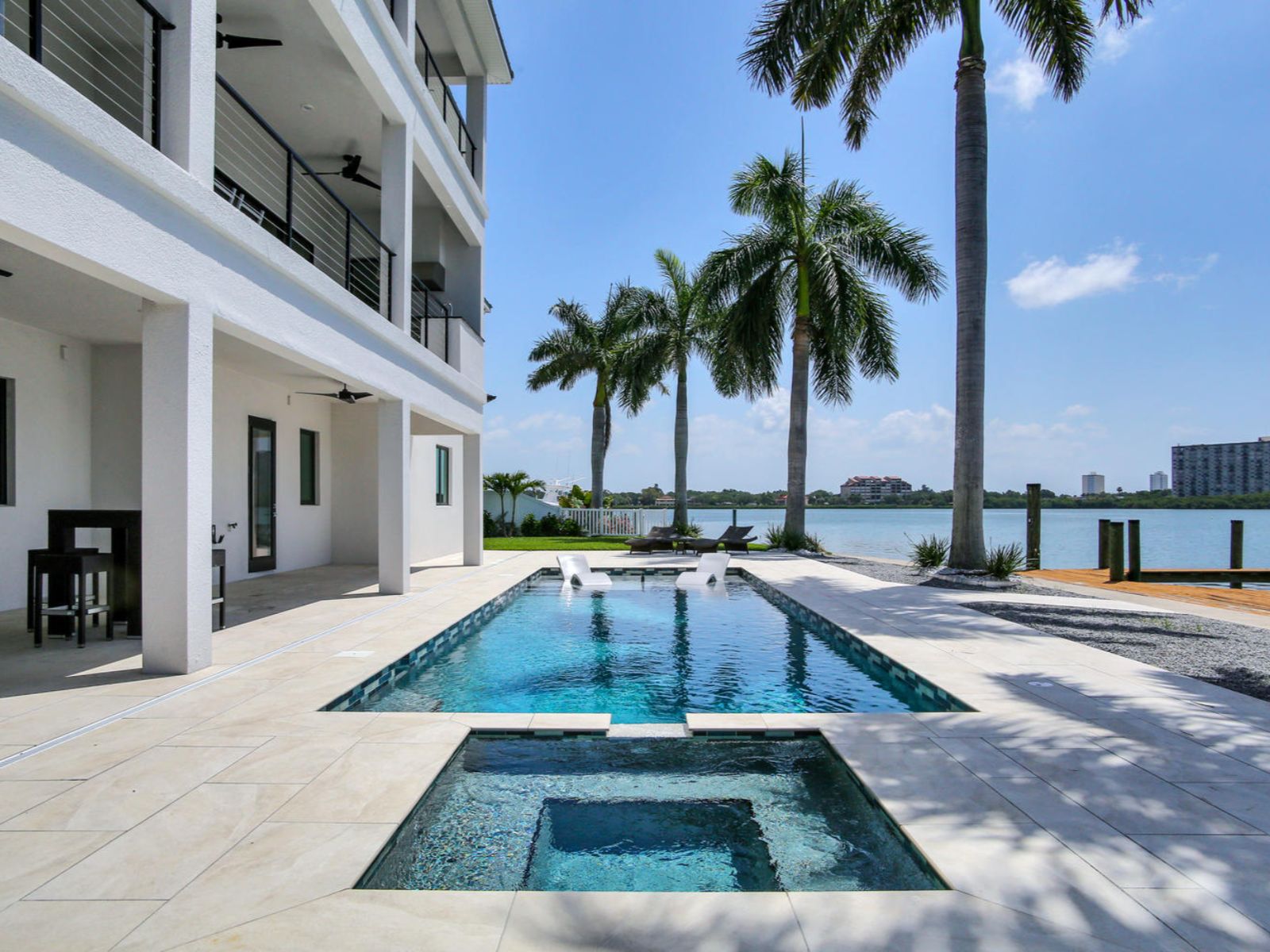 Modern waterfront property featuring a luxurious swimming pool and spa, surrounded by palm trees. The outdoor area includes a spacious patio with lounge chairs and a view of the serene waterway, ideal for relaxation and entertainment. Bright blue skies enhance the inviting atmosphere of this upscale retreat.