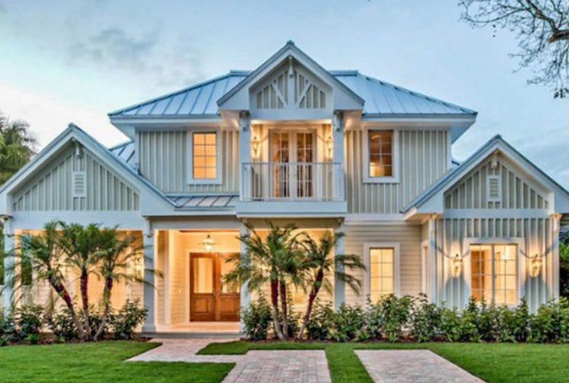 Modern coastal-style home with a light-colored exterior, featuring a metal roof, large windows, and a welcoming front porch surrounded by lush greenery and landscaping.