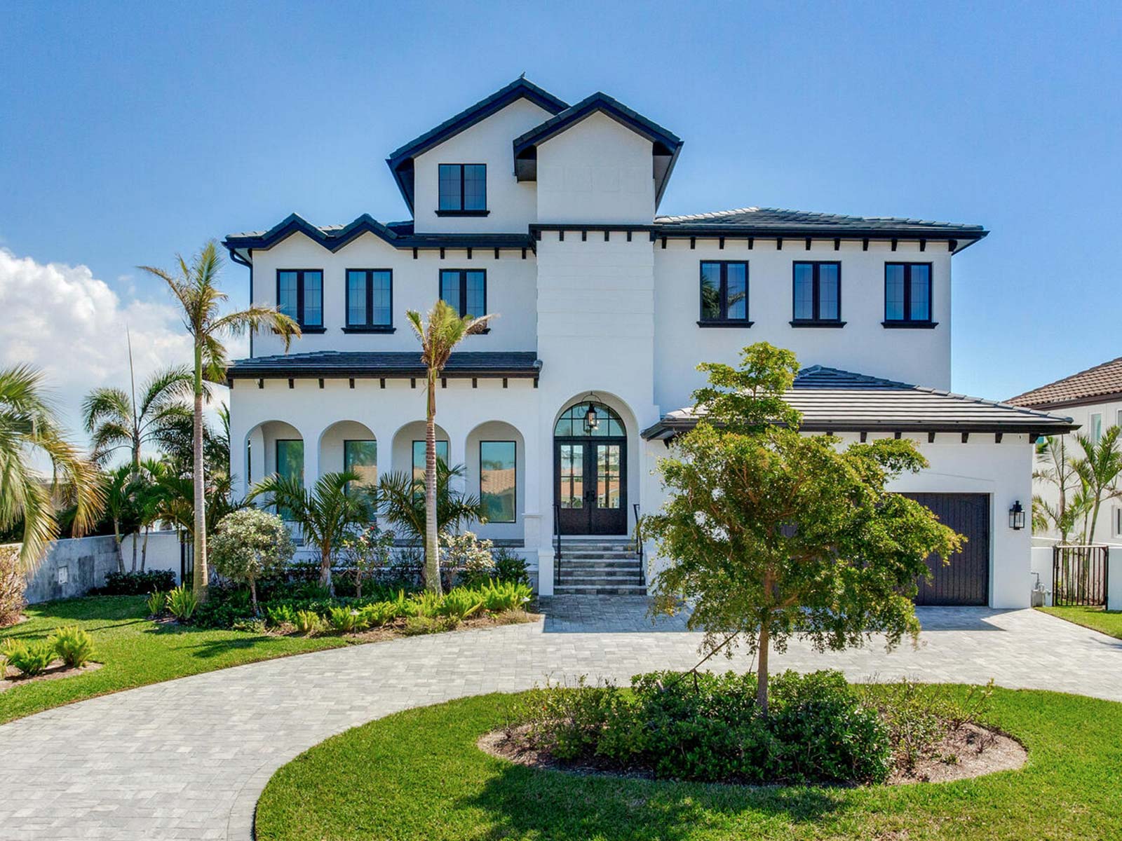 Modern two-story luxury home with a white exterior, black accents, and a landscaped front yard featuring palm trees and shrubs. The house has large windows, a decorative entrance, and a paved driveway.