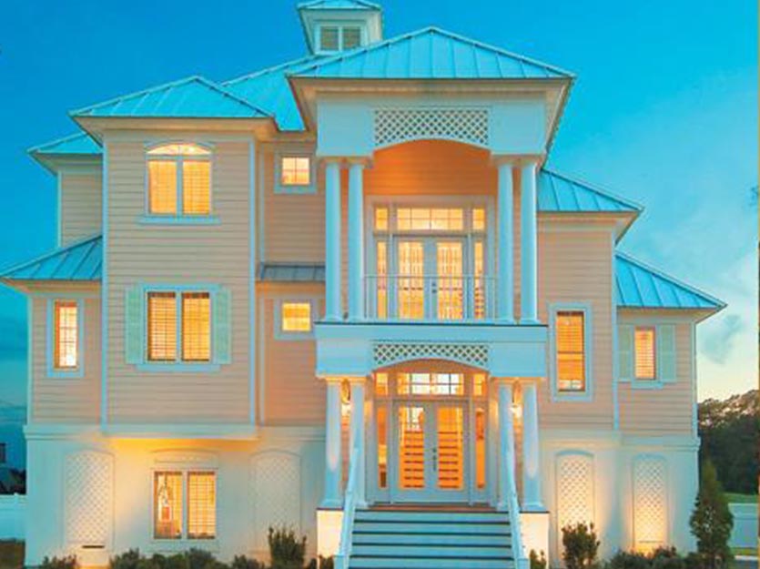 Elegant coastal-style home featuring a light-colored exterior, large windows, and a welcoming porch. The house showcases a modern design with a metal roof and detailed architectural elements, illuminated in the evening light. Ideal for beachside living and luxury home aesthetics.