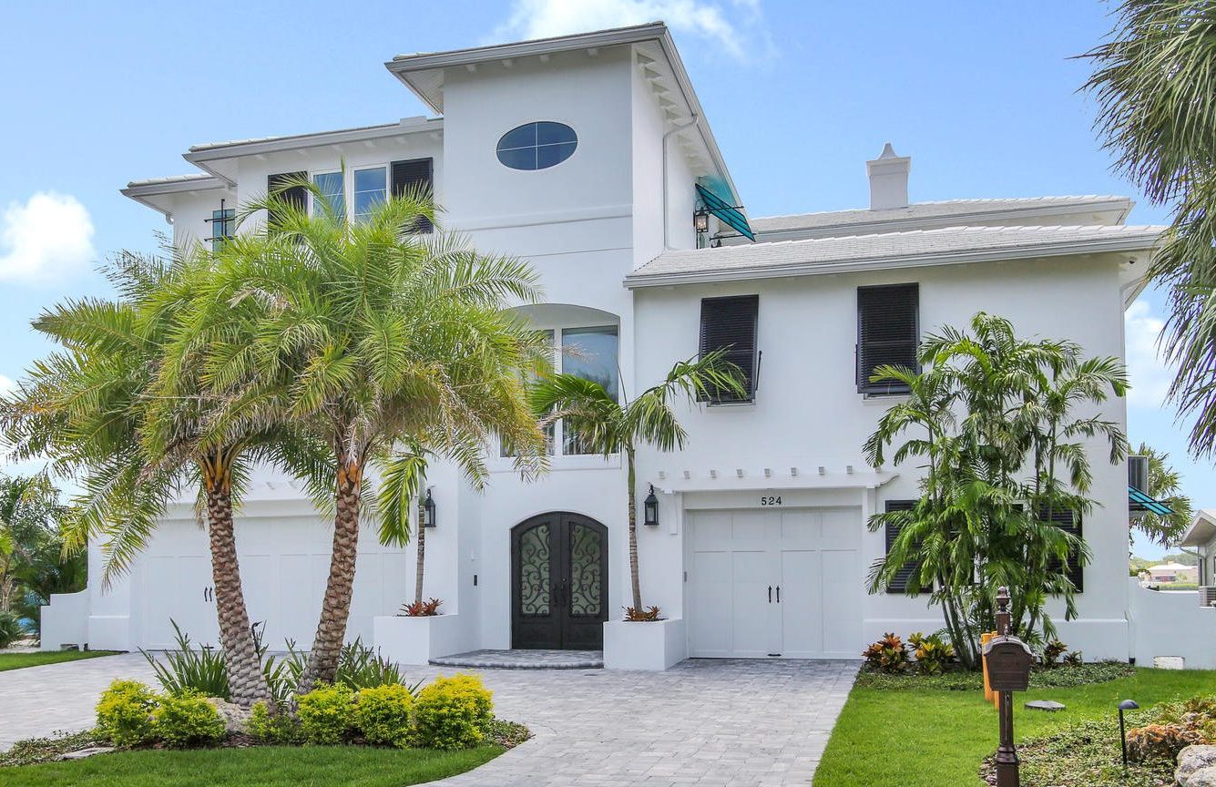 Modern white luxury home with palm trees and landscaped garden, featuring a double door entrance and a paved driveway.