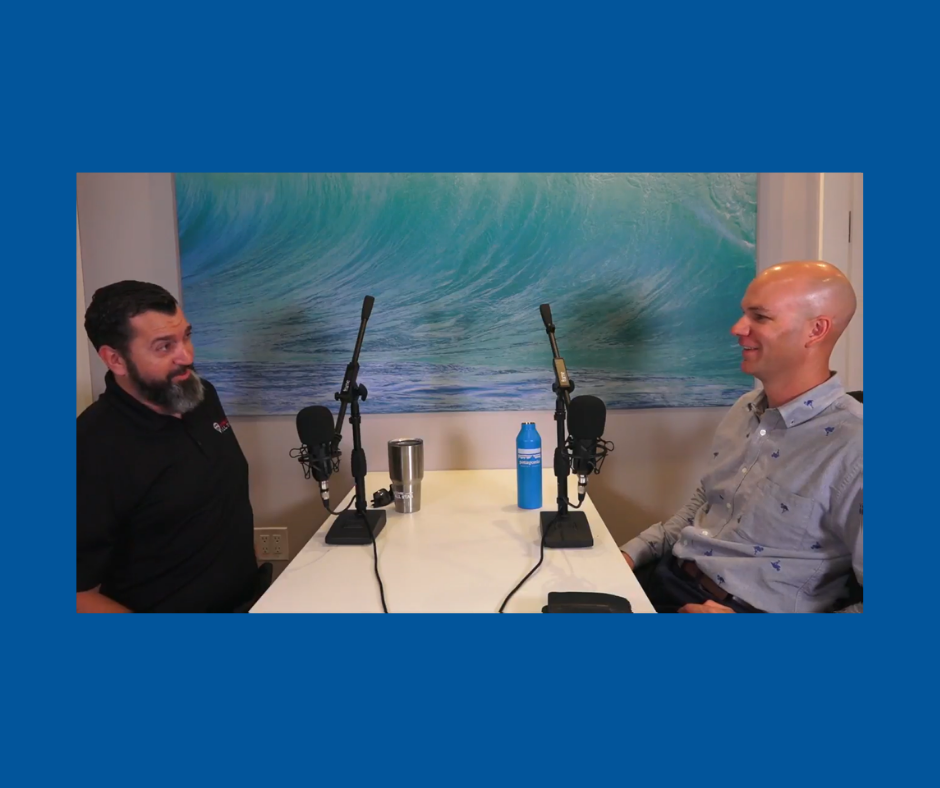 Two men engaged in a podcast discussion at a table with microphones, featuring a backdrop of ocean waves. One man has a beard and wears a black shirt, while the other has a bald head and a patterned button-up shirt. The table is equipped with a water bottle and a tumbler.