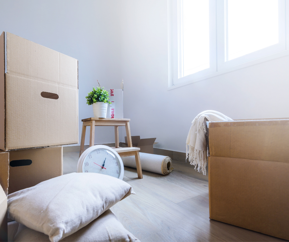 Moving boxes and decorative items in a bright, empty room, featuring a small plant on a wooden stool, a clock on the floor, and soft pillows stacked nearby. Ideal for home organization and moving inspiration.
