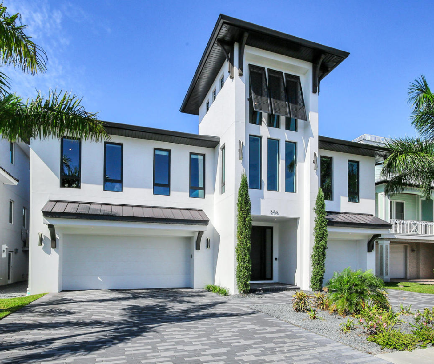 Modern luxury home with a white exterior, large windows, and a distinctive tower. Features a landscaped driveway with palm trees and vibrant greenery. Ideal for showcasing contemporary architectural design.