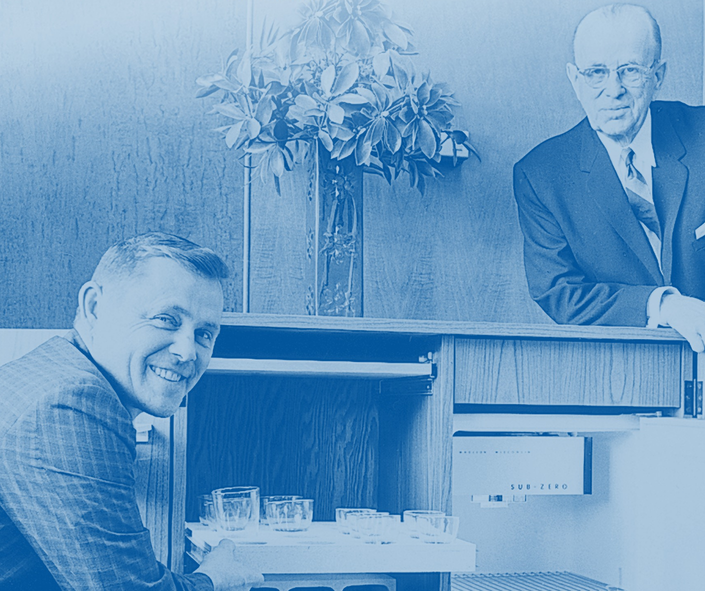 Two men in vintage attire are seen interacting with a Sub-Zero refrigerator, showcasing its features. One man is smiling while holding a tray with glasses, and the other is leaning against the refrigerator. A plant is visible in the background, adding to the homey atmosphere.