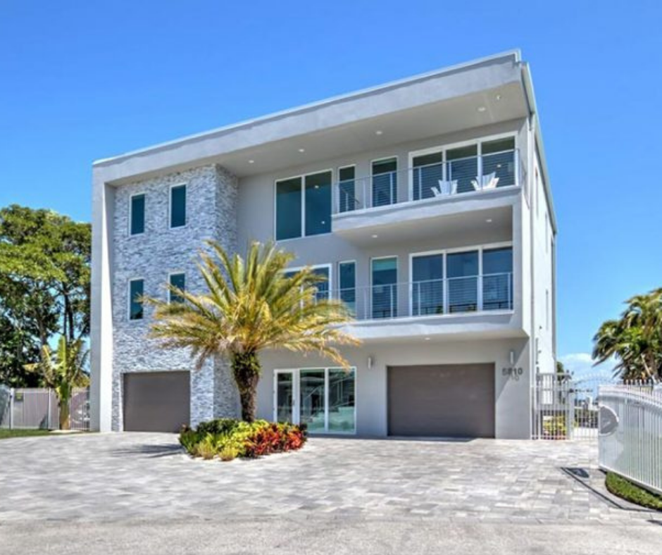 Modern three-story house with large windows, stone accents, and a landscaped front yard featuring palm trees and colorful plants, located in a sunny outdoor setting.