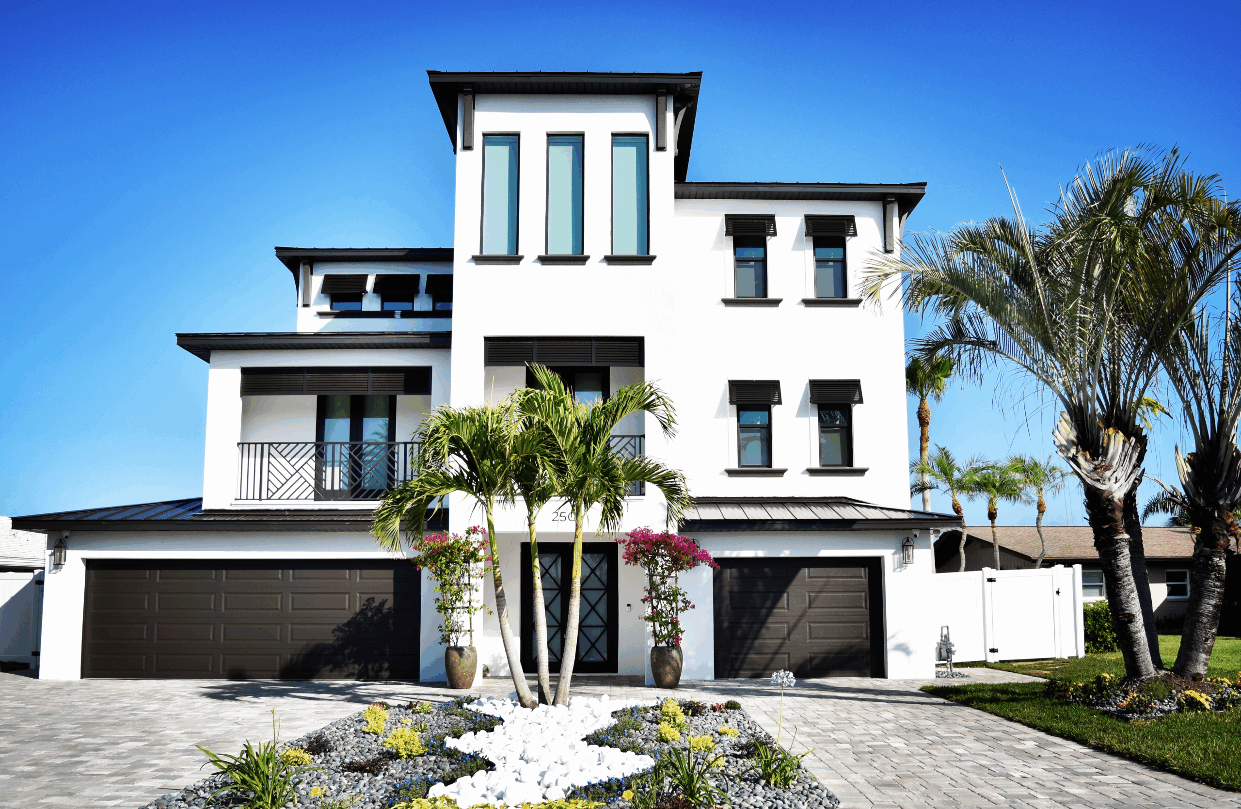Modern two-story white luxury home with a stylish facade, featuring large windows, a balcony, and tropical landscaping. The driveway is paved with stones, and palm trees and colorful flowers enhance the attractive curb appeal. Clear blue sky in the background.