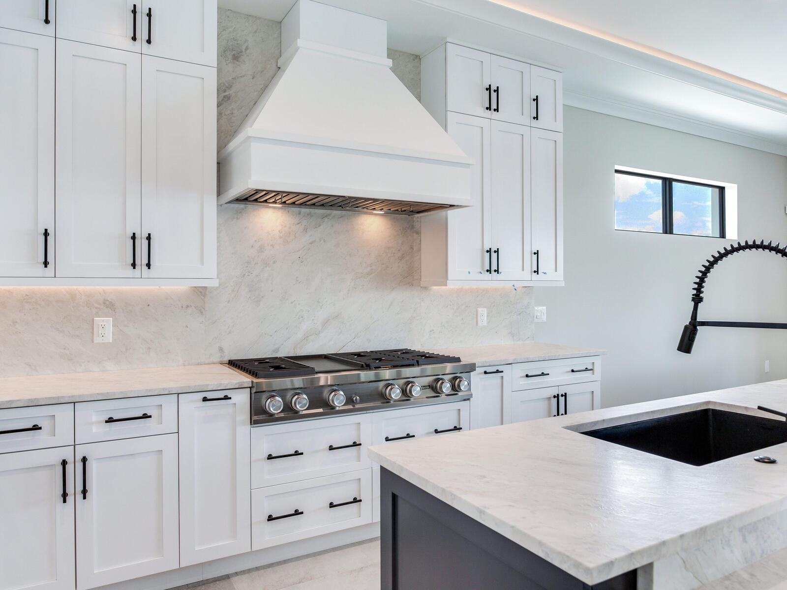 Modern kitchen with white cabinetry, a marble backsplash, and a stainless steel gas range. Features include black hardware, a sleek island with a black sink, and ample natural light from a window. Ideal for contemporary home design inspiration.