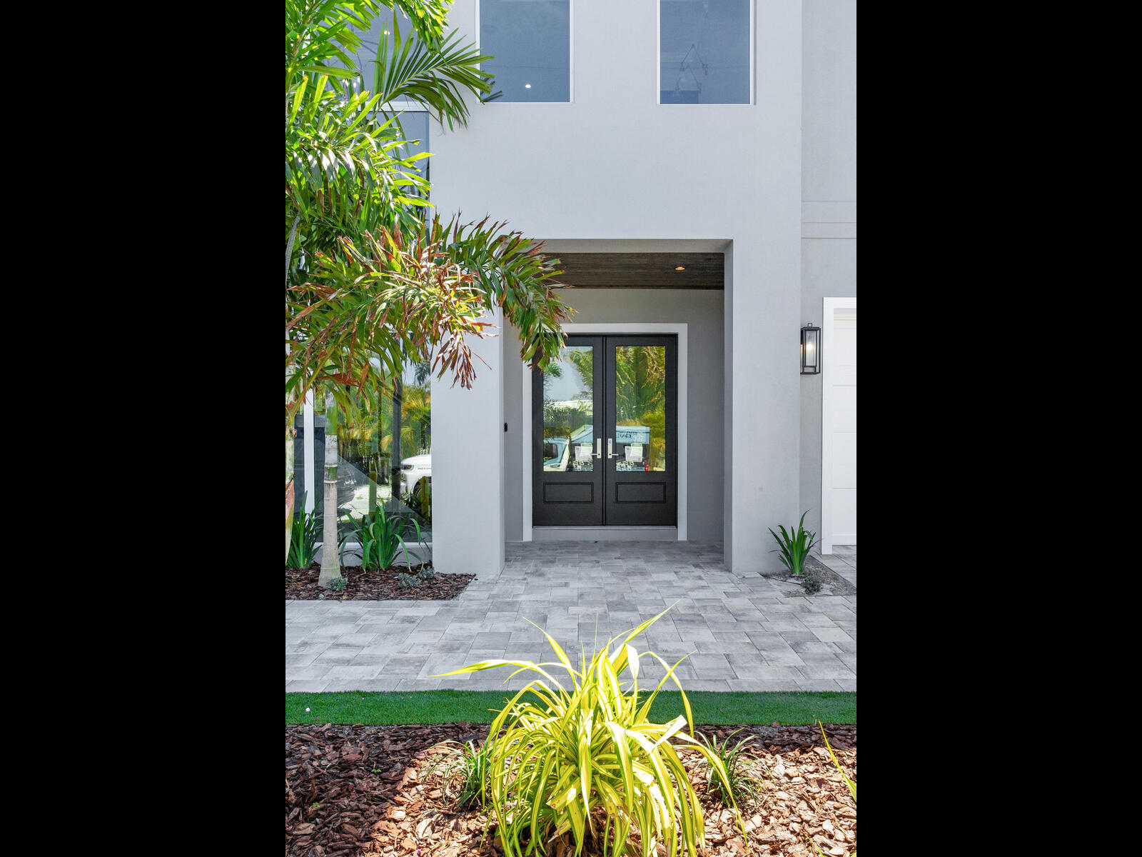 Modern home entrance featuring double doors, surrounded by lush greenery and landscaped pathways. The exterior showcases a contemporary design with a clean, gray facade and stylish lighting.