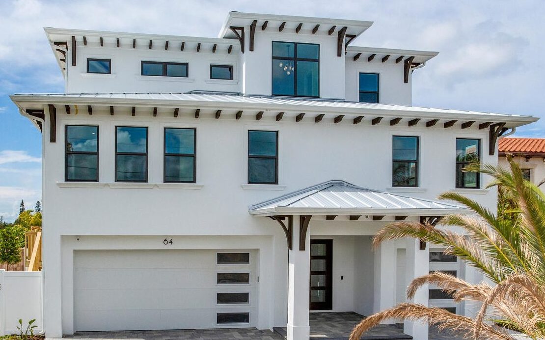 Modern two-story white house with large windows and a metal roof, featuring a spacious front porch and a garage, surrounded by palm trees. Ideal for showcasing contemporary architecture.