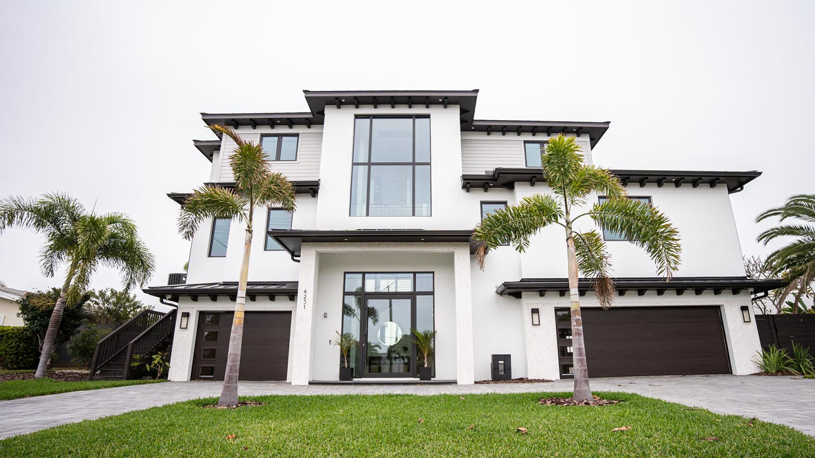 Modern two-story house with large windows and a sleek design, featuring a manicured lawn, palm trees, and a paved driveway. The exterior showcases a combination of white and dark elements, highlighting contemporary architecture.