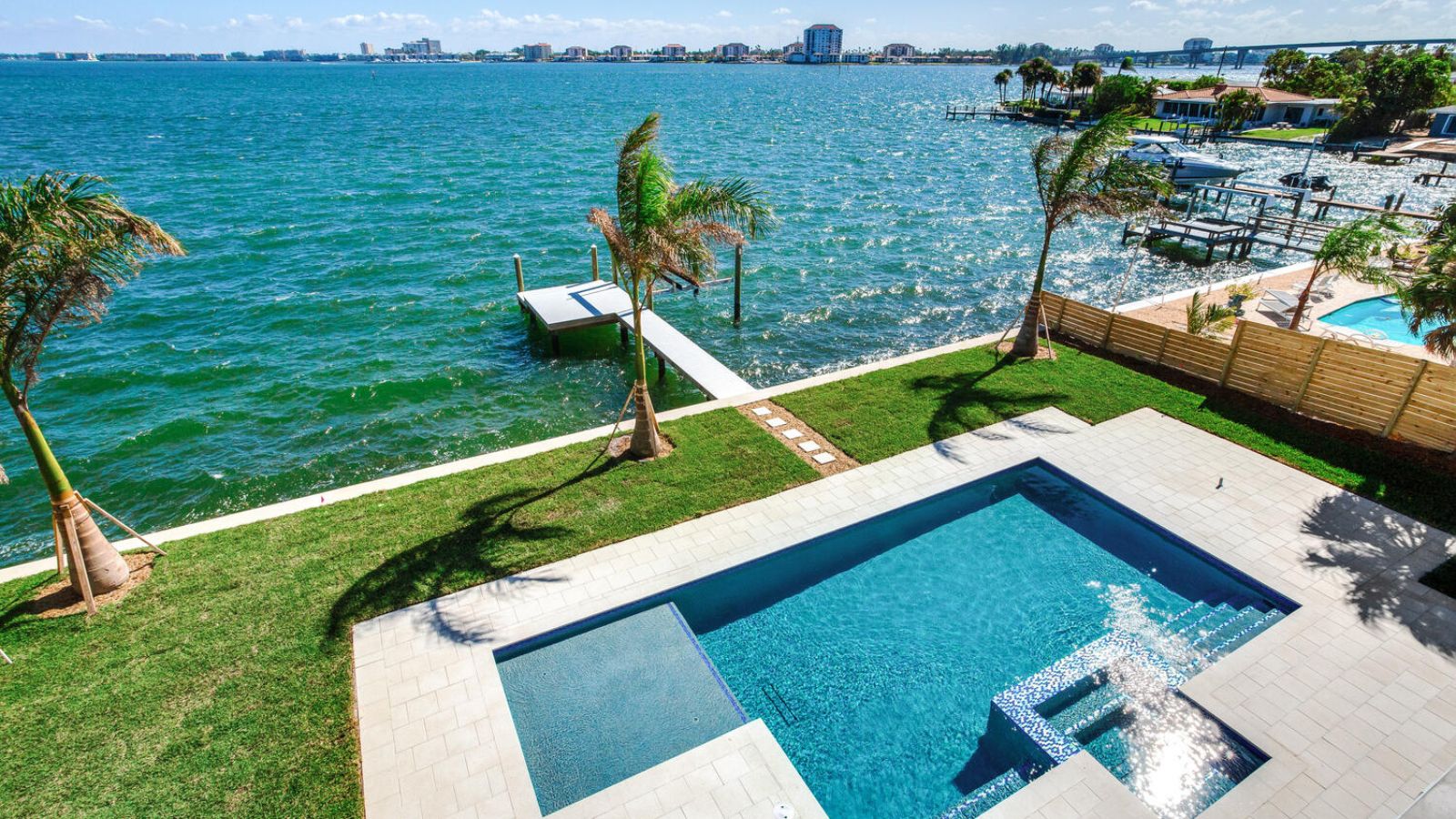 Scenic waterfront view featuring a luxurious pool, lush green lawn, and a private dock overlooking vibrant blue water and a distant shoreline with buildings under a clear sky.
