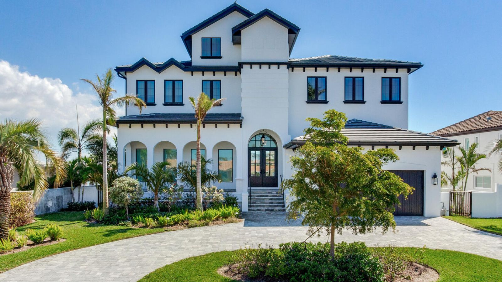 Modern two-story white house with a black roof, featuring large windows, a welcoming front entrance, and landscaped greenery, including palm trees and shrubs, set against a clear blue sky.