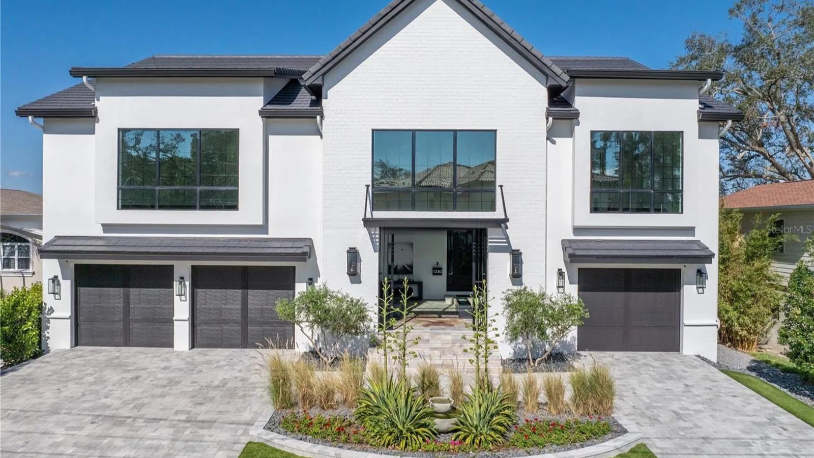 Modern two-story white brick house with large windows, featuring a landscaped front yard, stone pathway, and double garage doors, set against a clear blue sky.