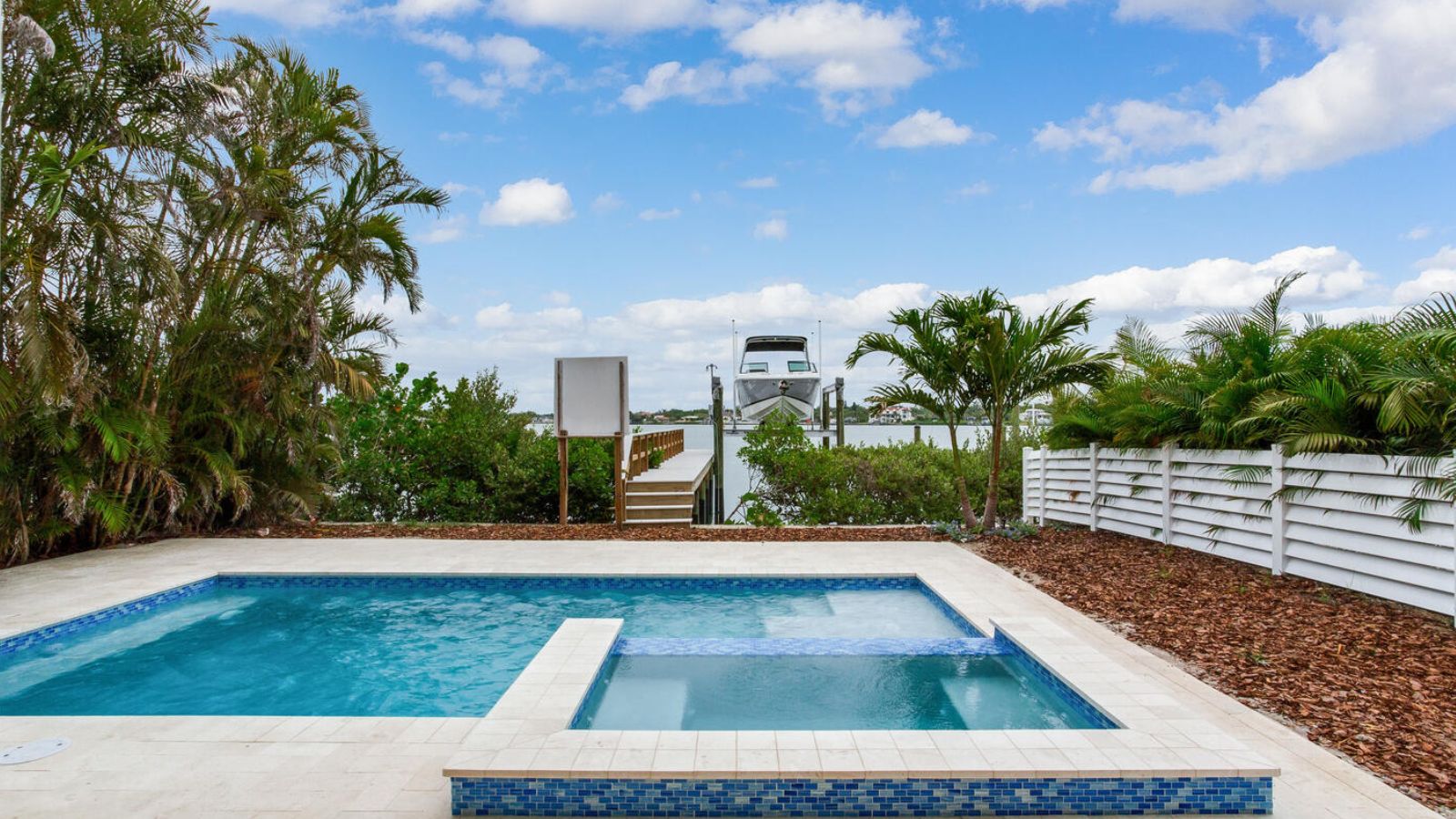 Outdoor pool area with a spa, surrounded by tropical foliage, overlooking a docked boat and waterfront landscape under a blue sky with clouds.