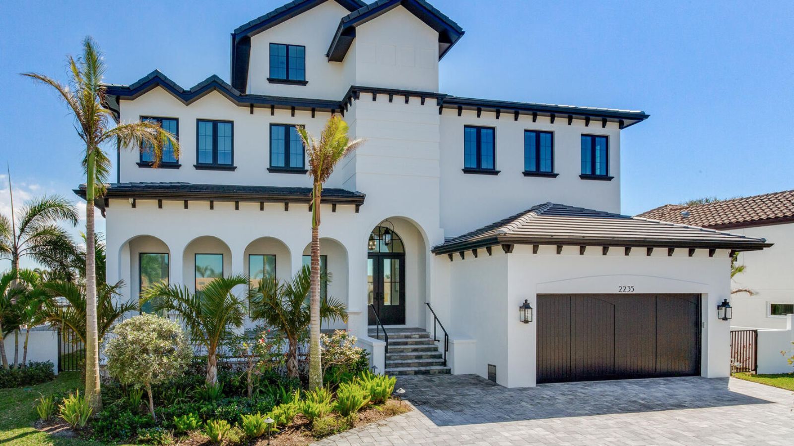 Modern two-story house with a white exterior, black trim, and a spacious driveway surrounded by lush landscaping and palm trees. The home features large windows and a stylish entrance with arched doorways, showcasing contemporary architectural design.