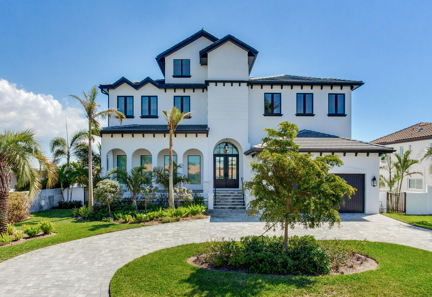 Modern two-story luxury home with a white exterior, black accents, and a landscaped front yard featuring palm trees and shrubs. The home has large windows and a stylish entrance with steps leading to the front door, surrounded by a circular driveway.