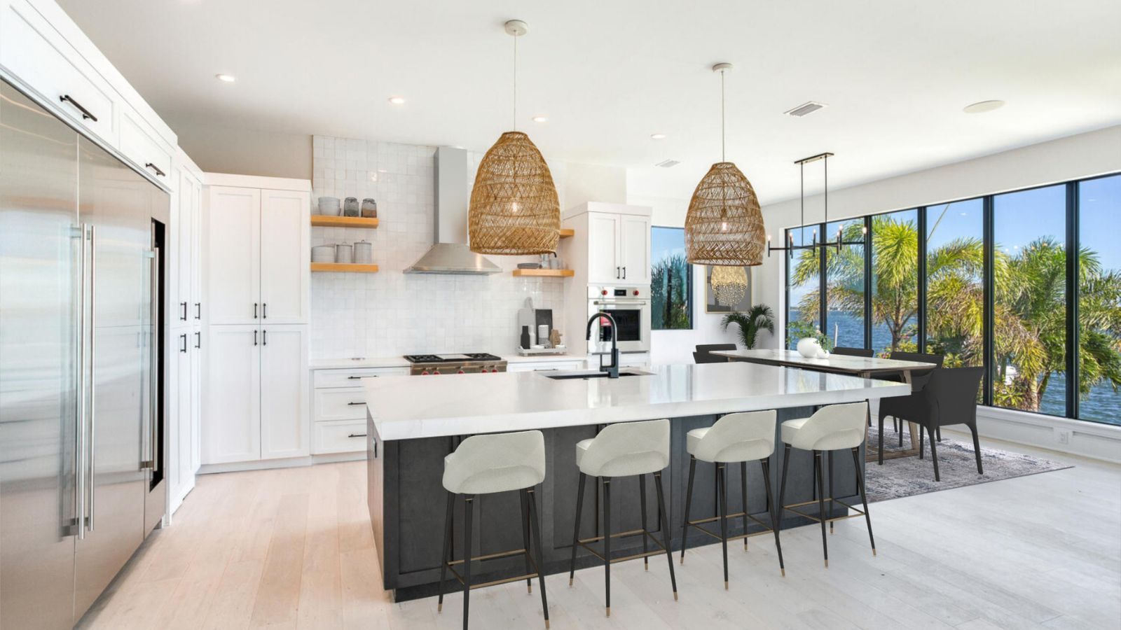 Modern kitchen featuring a large island with seating, sleek white cabinetry, stainless steel appliances, and stylish woven pendant lights. The space is bright and airy, with large windows showcasing a view of palm trees and water.