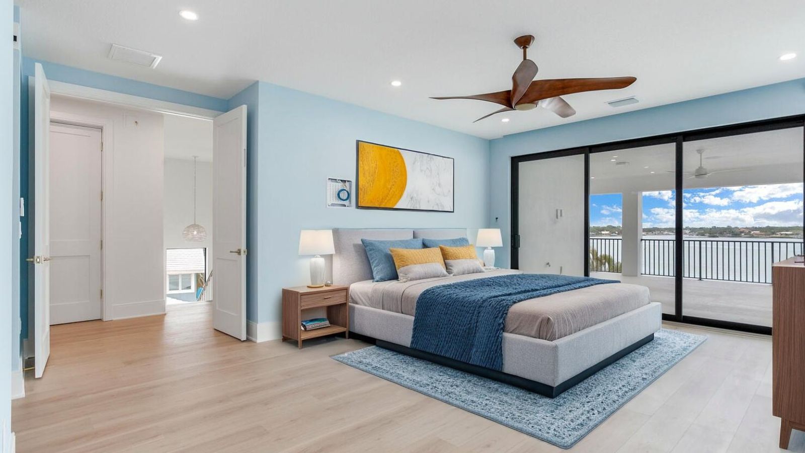 Modern bedroom featuring a queen-sized bed with blue and gray bedding, stylish decor, and large sliding glass doors that open to a balcony with a water view. The room has light blue walls, wooden flooring, and contemporary lighting fixtures, creating a serene and inviting atmosphere.