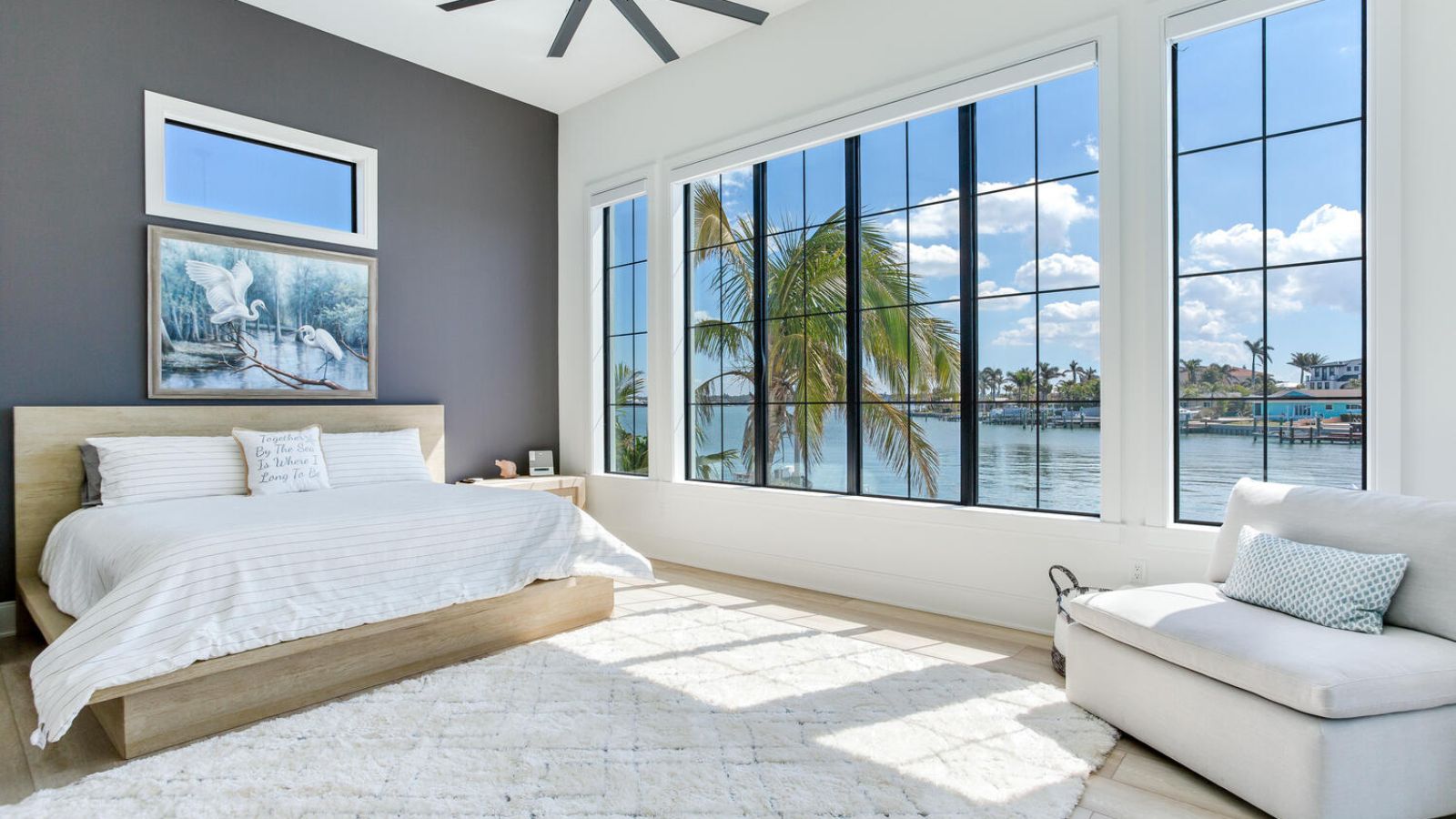 Bright and airy bedroom featuring a modern design with a wooden bed, decorative pillows, and a cozy rug. Large windows offer a stunning view of the waterfront and palm trees, enhancing the serene atmosphere. Ideal for relaxation and coastal living.