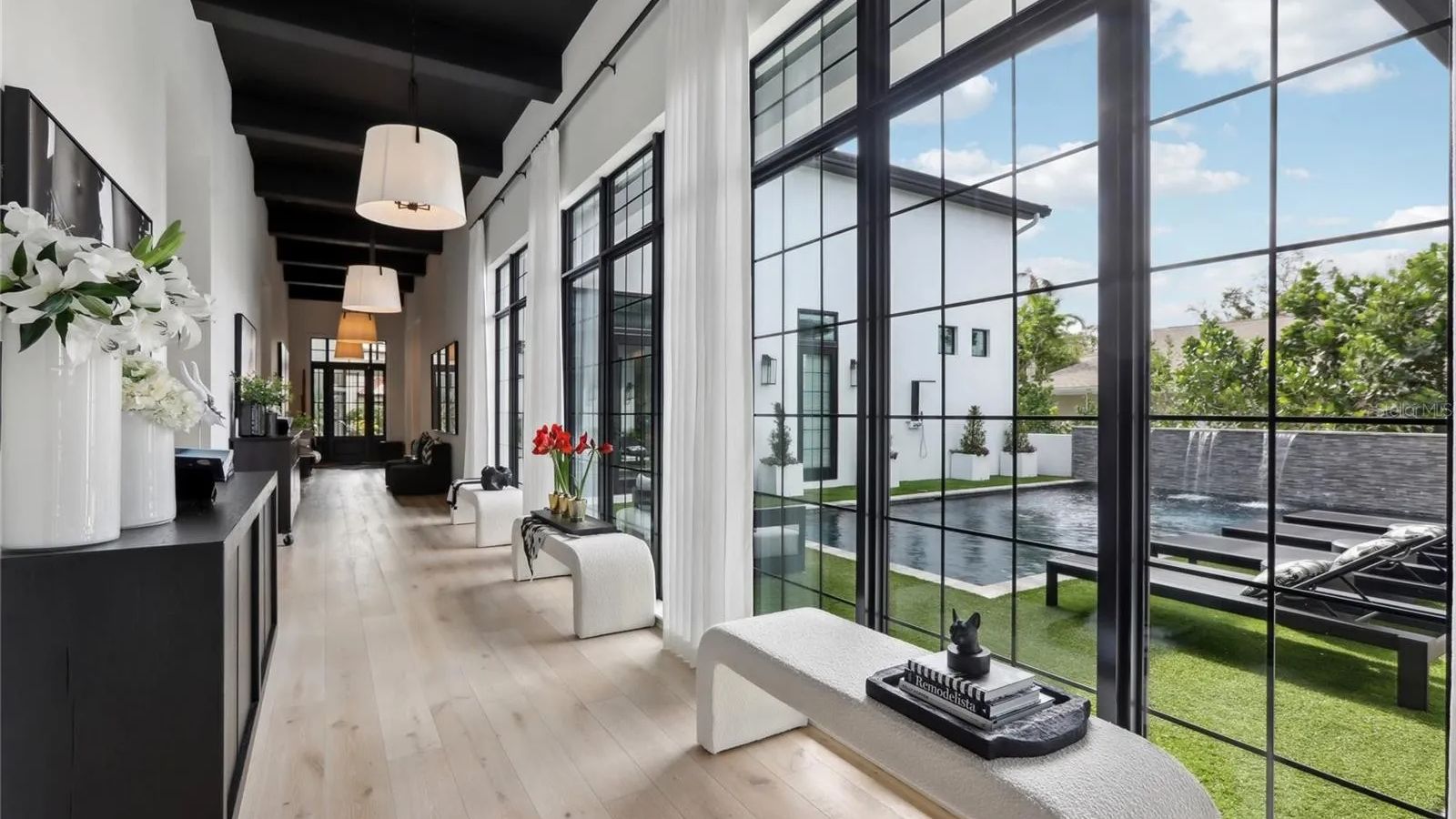 Modern hallway featuring large windows with black frames, natural light illuminating light wood flooring, stylish white and black decor, and a view of a serene outdoor pool area with greenery.