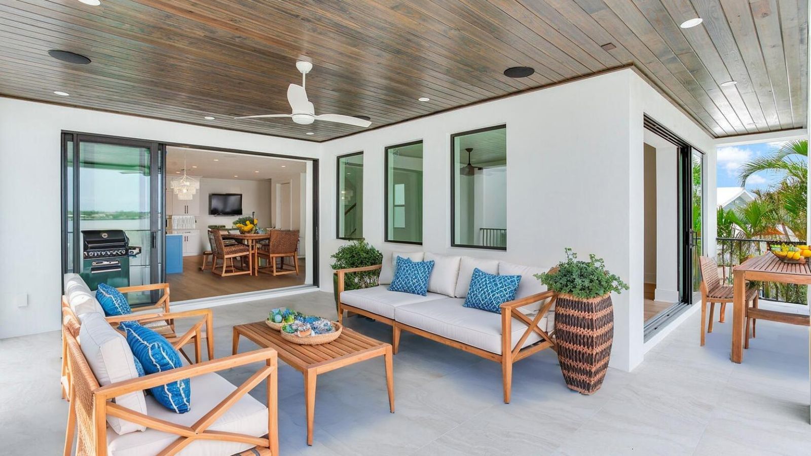 Modern outdoor living space featuring comfortable seating with blue accent pillows, a wooden coffee table, and lush greenery. The space opens up to an indoor dining area with a view, complete with a television and a stylish chandelier. A barbecue grill is visible, enhancing the entertaining potential of this bright, airy patio.