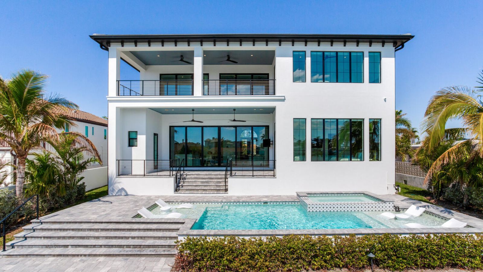 Modern luxury home with a spacious backyard pool area, featuring palm trees and a sleek outdoor design, ideal for relaxation and entertainment. The house boasts large windows, multiple balconies, and contemporary architecture under a clear blue sky.