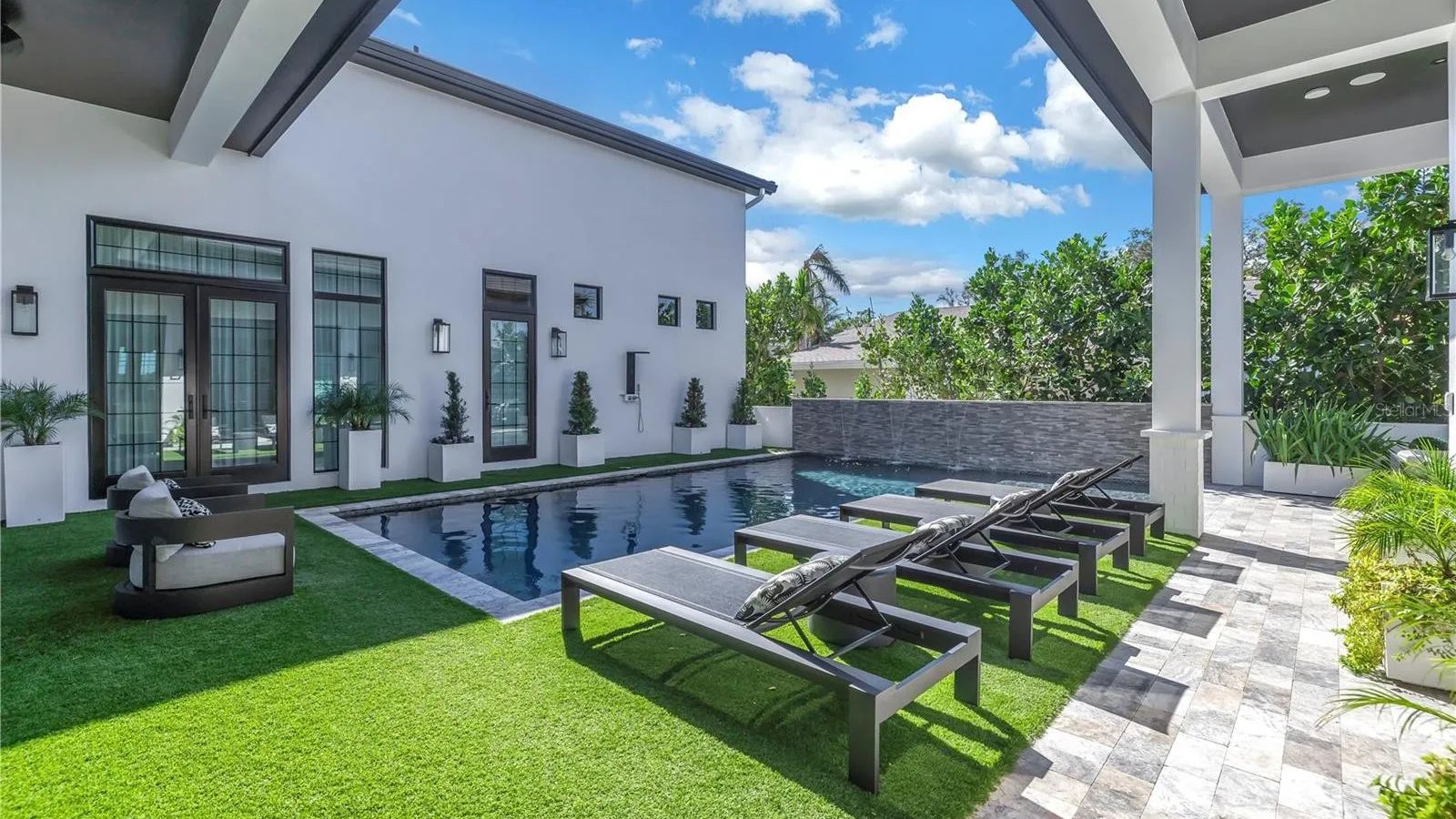 Modern outdoor space featuring a luxurious pool, comfortable lounge chairs, and stylish landscaping. The area is designed for relaxation, with artificial grass and decorative plants enhancing the aesthetic appeal. Bright blue skies and fluffy clouds complete the serene atmosphere.