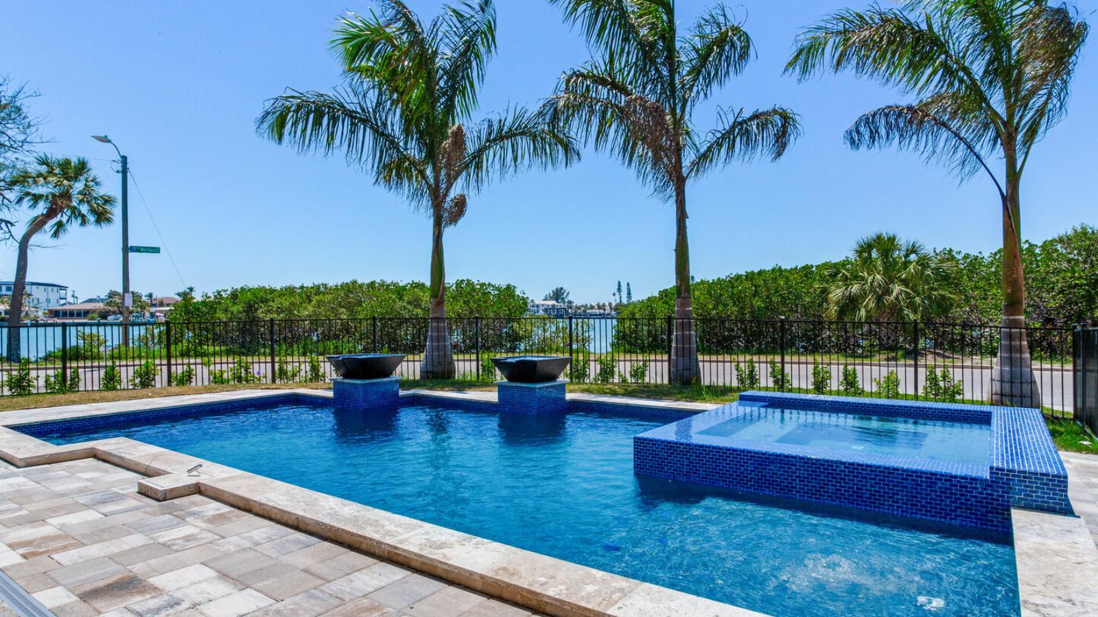 waterfront swimming pool with a raised spa, blue mosaic tile, fire bowls on pedestals, paver decking, palm trees, and a view of the bay