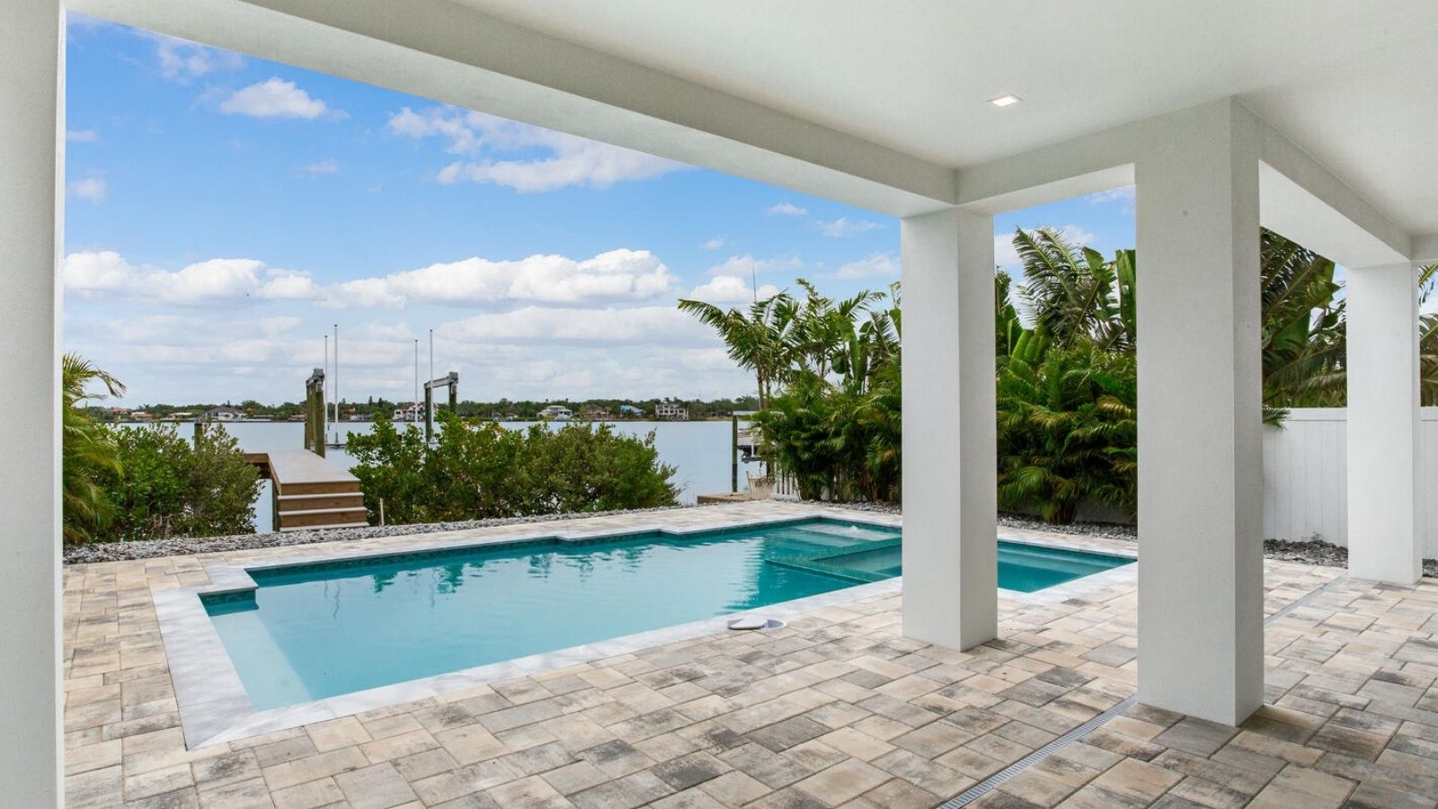 covered patio with swimming pool and spa, waterfront view of a bay and boat dock, surrounded by tropical landscaping and paver decking.