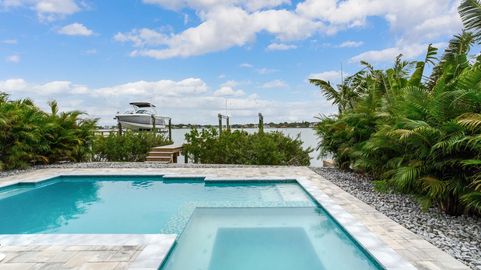 waterfront swimming pool with integrated spa, marble coping, gravel border, tropical landscaping, and a boat lift on the intercoastal waterway