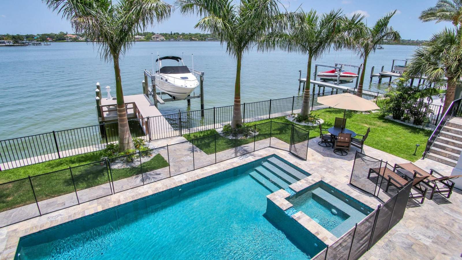 Waterfront backyard featuring a swimming pool, surrounded by a black fence, with a boat lift and dock visible on the water