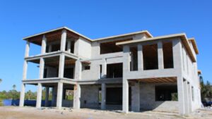 Newly constructed multi-story building with a concrete block exterior, featuring an unfinished facade and exposed beams under a clear blue sky. The site shows signs of ongoing construction, with debris and equipment scattered around the perimeter.