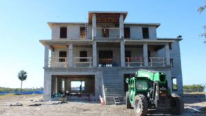 Construction site of a multi-story building featuring concrete structure, scaffolding, and a green telehandler, with palm trees and clear blue sky in the background.