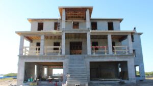 Construction site of a multi-story concrete house with unfinished exterior, featuring a staircase leading to the entrance, scaffolding, and construction materials visible around the site.