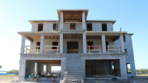 Construction site of a multi-story concrete house with exposed framework and stairs, showcasing ongoing building work under a clear blue sky.