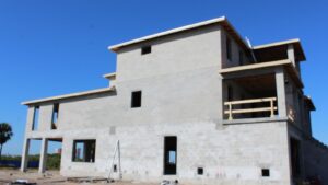 Newly constructed two-story building made of concrete blocks, showcasing a modern architectural design with open windows and unfinished exterior. The construction site is clear of debris, set against a blue sky.