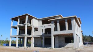 Construction site of a multi-story concrete house, showcasing unfinished exterior walls and exposed framing under a clear blue sky.