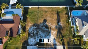 Aerial view of a vacant waterfront lot surrounded by residential properties with a pool, palm trees, and parked vehicles, showcasing the potential for new construction in a scenic neighborhood.