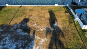 Aerial view of a grassy waterfront property with shadows cast by nearby trees, showcasing a clear blue waterway and a swimming pool in the background.