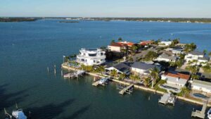 Aerial view of a waterfront property featuring a modern white house with a circular design, surrounded by palm trees and a marina, with boats docked along the shoreline and residential homes in the background. The scene captures the serene blue water and clear sky, highlighting a picturesque coastal neighborhood.