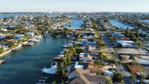 Aerial view of a residential waterfront community featuring houses along canals, boats docked at private piers, and lush palm trees, with a skyline visible in the distance.