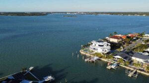 Aerial view of waterfront homes along a serene bay, featuring a modern white house with a round design and private dock, surrounded by palm trees and boats, showcasing the tranquil beauty of coastal living.