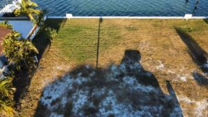 Aerial view of a waterfront property with a grassy yard bordered by palm trees, showcasing a shadow cast by a nearby structure, and the shimmering water in the background.