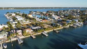 Aerial view of a coastal residential area featuring waterfront homes, docks, and boats along the calm blue waters, surrounded by palm trees and greenery in a sunny setting.