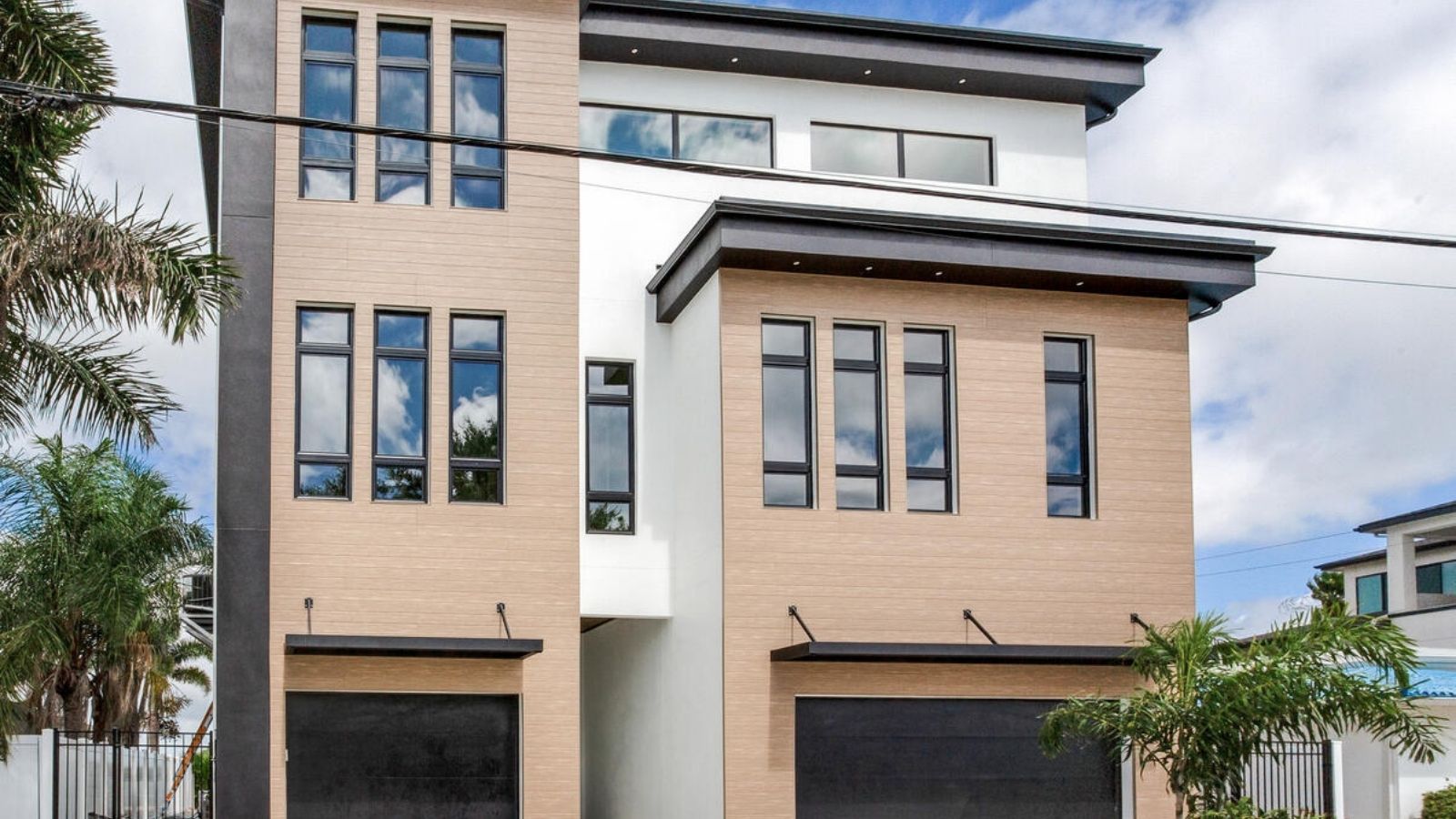 Modern three-story residential building featuring a combination of wood and sleek black accents, large windows, and two garage doors, surrounded by tropical landscaping.